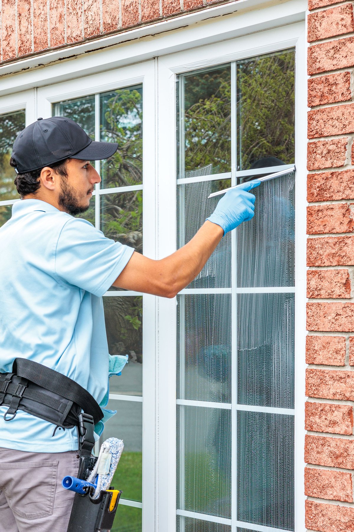 Professional window cleaner in blue shirt cleaning a large residential window with blue gloves and tool belt