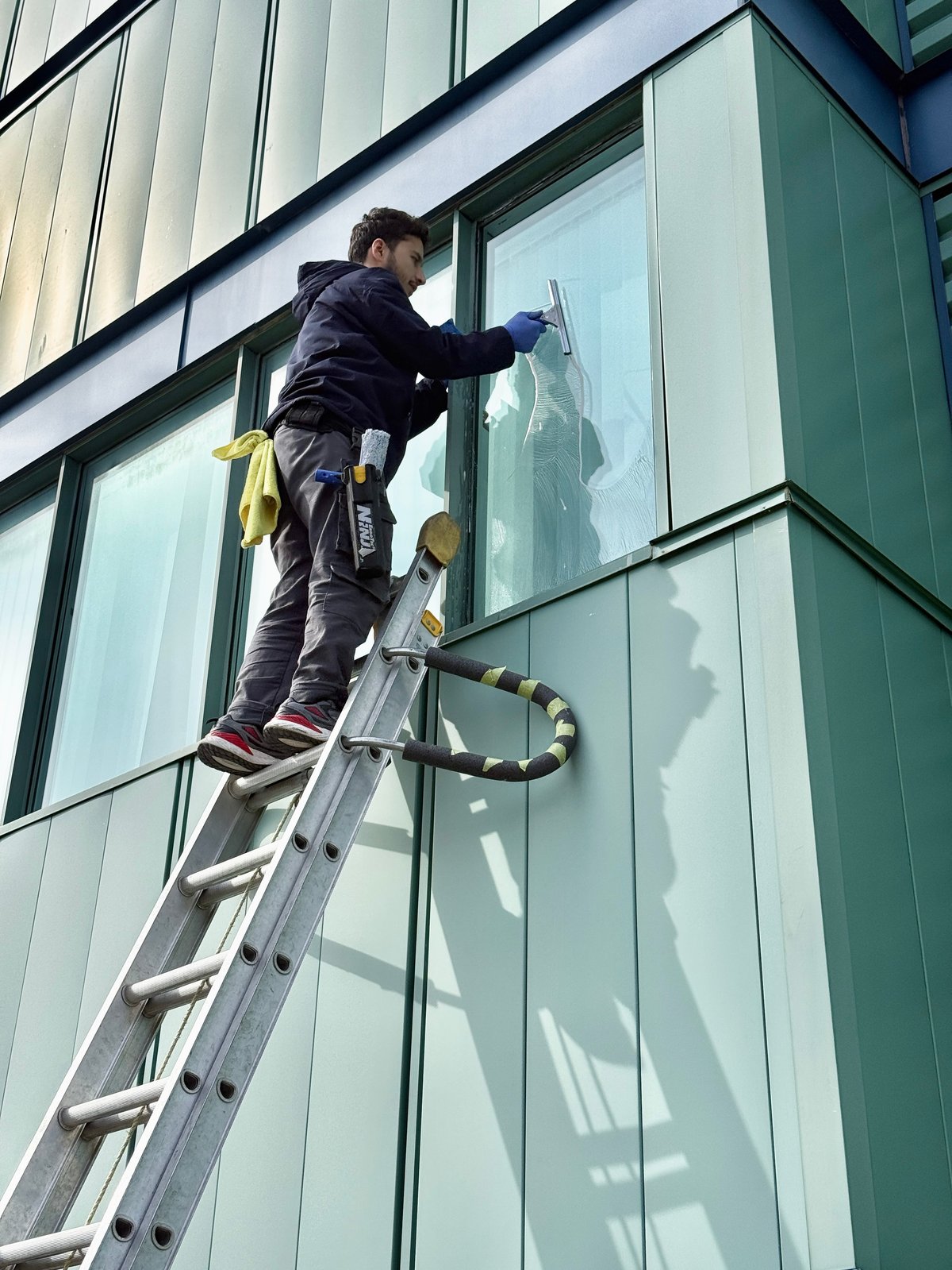 Worker on ladder cleaning large building windows with professional equipment and safety gear