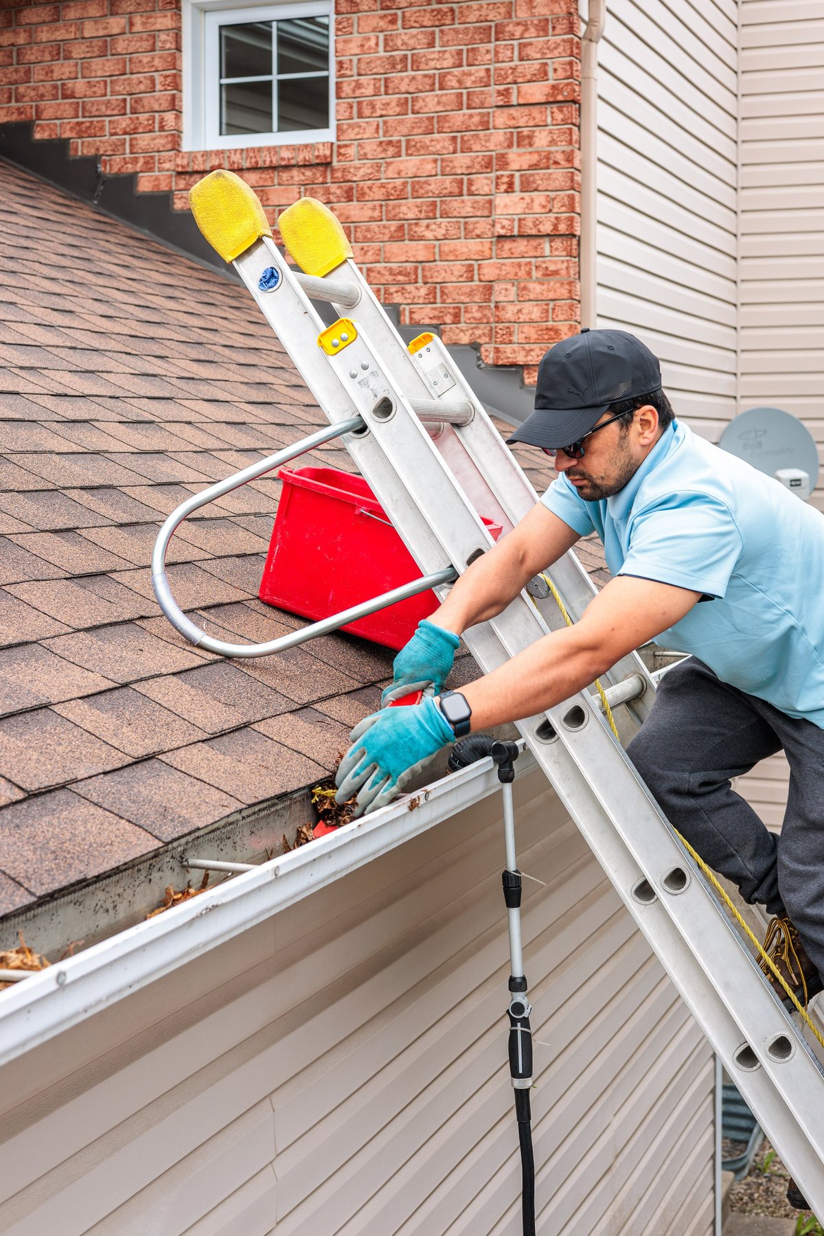 Worker cleaning house gutter with ladder against brick home and siding